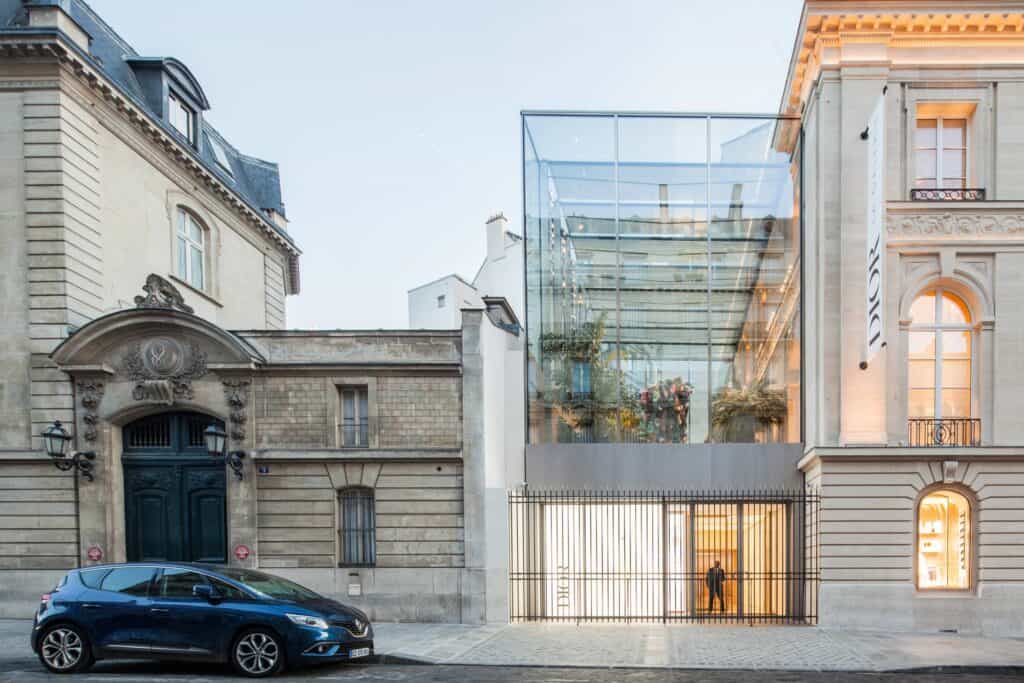 New glass and steel winter garden extension of Dior 30 Montaigne on rue François Ier, set between two carved stone hôtels particuliers, with plants behind the glazing and a gated entrance below