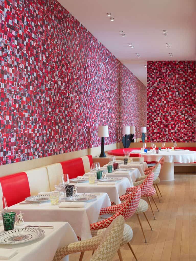 Monsieur Dior restaurant at Dior 30 Montaigne, white-clothed tables, red banquettes and red-and-white patterned chairs along a wall covered in a vivid red and pink collage of archival photographs