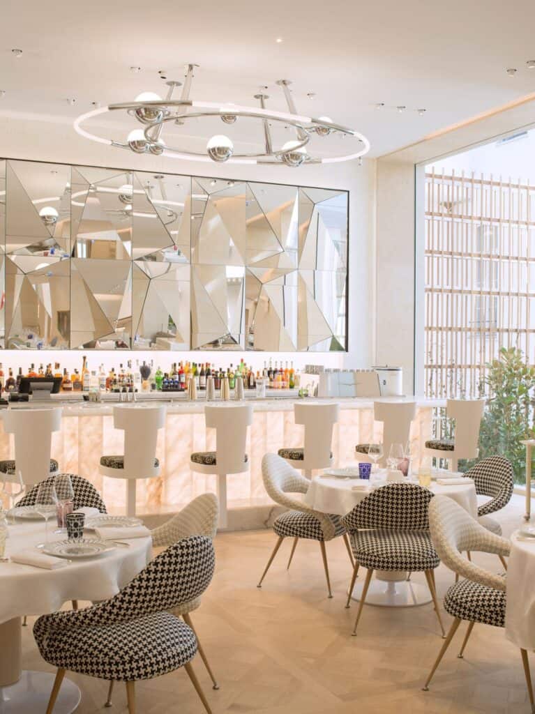 Bar at Dior 30 Montaigne with a backlit stone counter, a faceted mirrored wall behind shelves of bottles, a sculptural silver chandelier and houndstooth-upholstered chairs at round tables