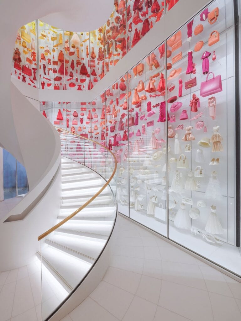 Colorama staircase in La Galerie Dior at Dior 30 Montaigne, a white spiral stair winding past a glass wall of miniature dresses, bags and shoes graded from red and pink to white