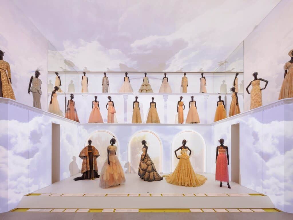 Room in La Galerie Dior at Dior 30 Montaigne with rows of gowns in gold, orange and red on mannequins on tiered white platforms, under a ceiling and walls painted with soft clouds