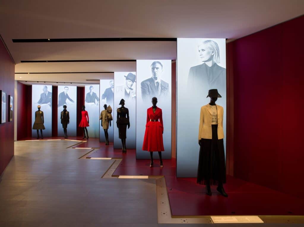 Red-walled gallery in La Galerie Dior at Dior 30 Montaigne with mannequins in Dior looks standing before tall illuminated black-and-white portraits of the house's creative directors