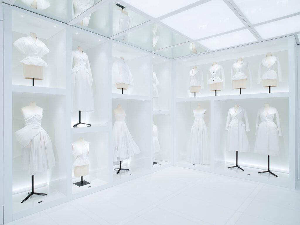 All-white room in La Galerie Dior at Dior 30 Montaigne with illuminated glass cases of white toiles and dresses on stands, beneath a glowing grid ceiling