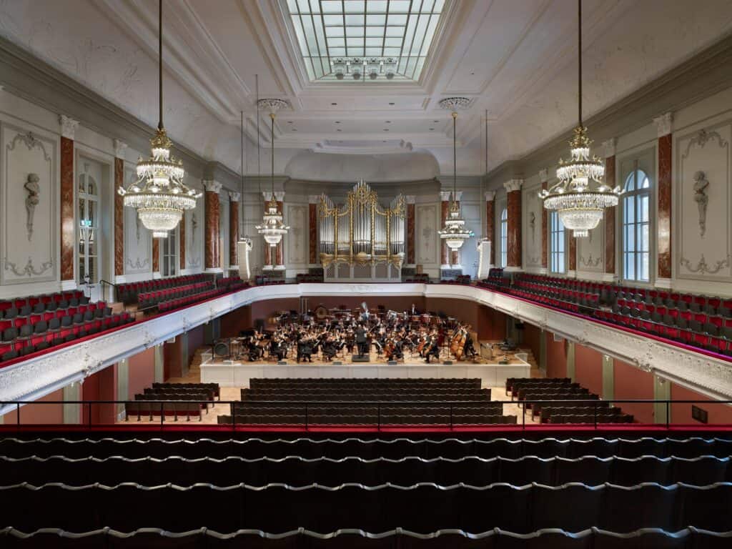 Musiksaal of the Stadtcasino Basel seen from the balcony with an orchestra playing on the stage, crystal chandeliers hanging from the white stucco ceiling and reopened skylight, marbled red columns and the gilded organ case behind