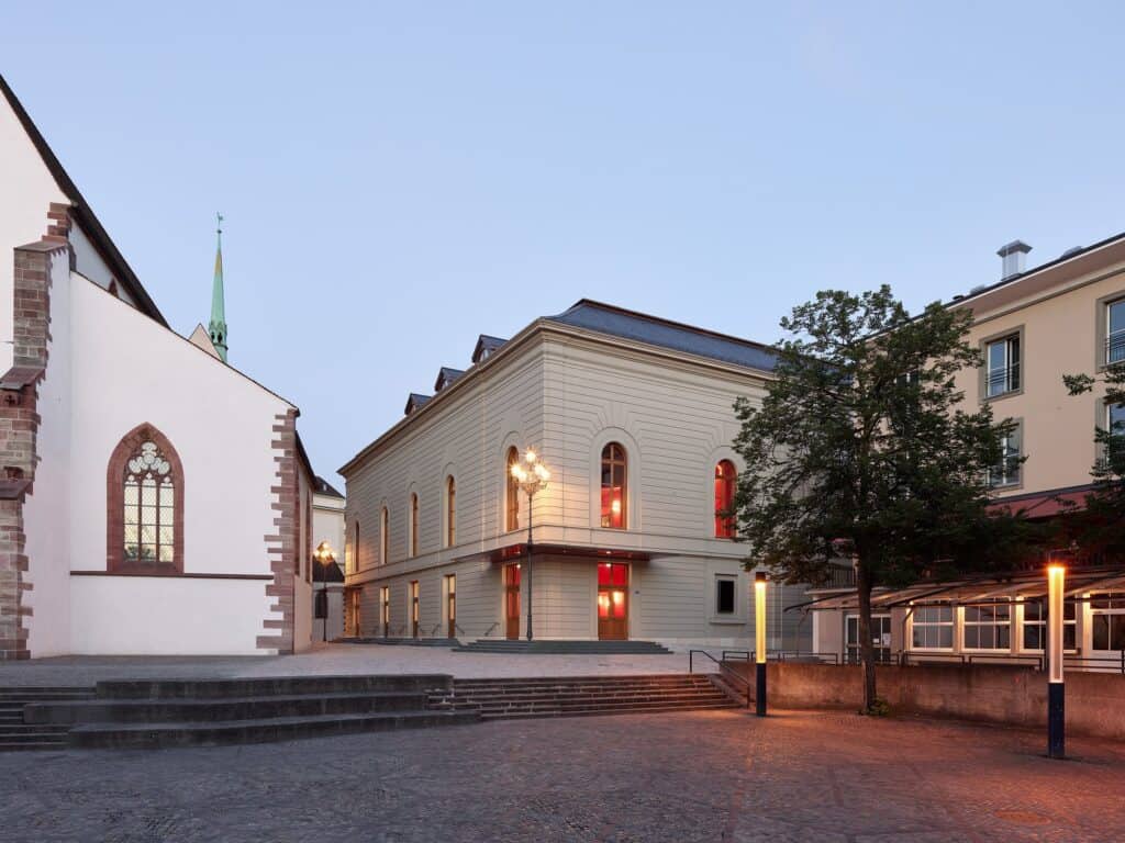 Stadtcasino Basel extension at dusk beside the Barfüsserkirche, its pale neo-Baroque facade of arched windows glowing red inside, a lantern-topped lamp post and broad steps on the cobbled square
