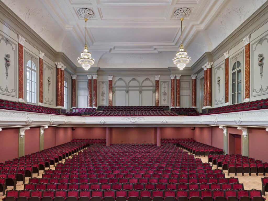 Musiksaal of the Stadtcasino Basel viewed from the stage over rows of red velvet seats to the rear balcony, marbled red pilasters, white stucco busts and two crystal chandeliers beneath the ornate white ceiling