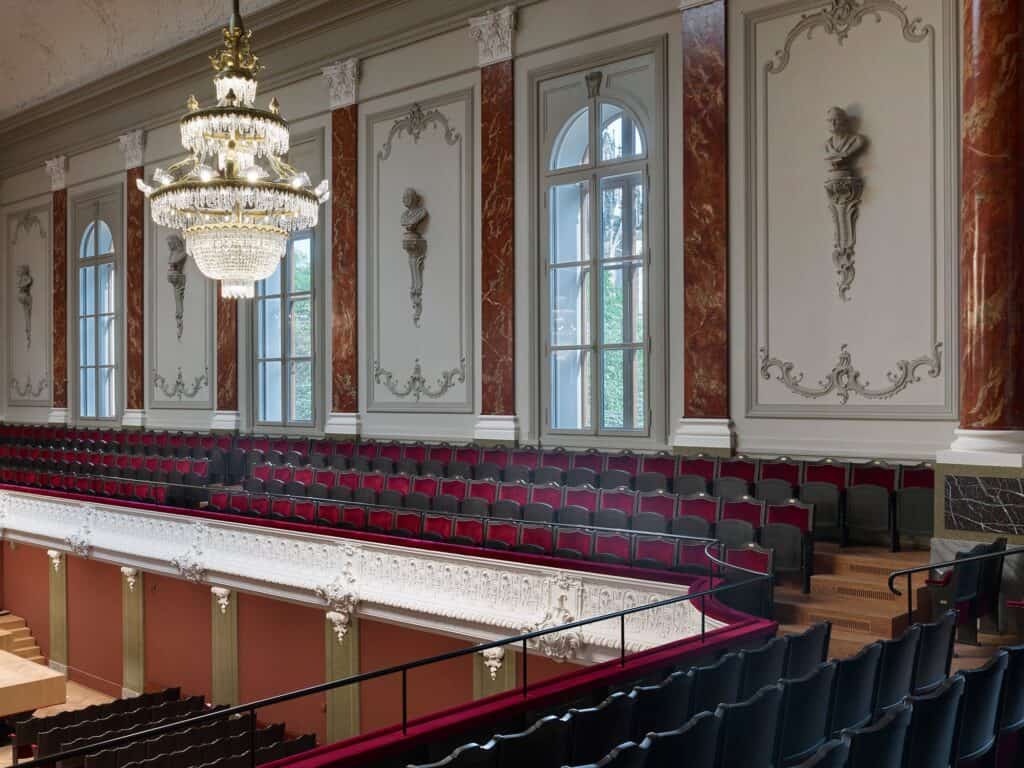 Side balcony of the Musiksaal at the Stadtcasino Basel with red seats behind a white stucco balustrade, marbled red pilasters, tall arched windows, a crystal chandelier and composer busts framed by stucco garlands on the grey walls