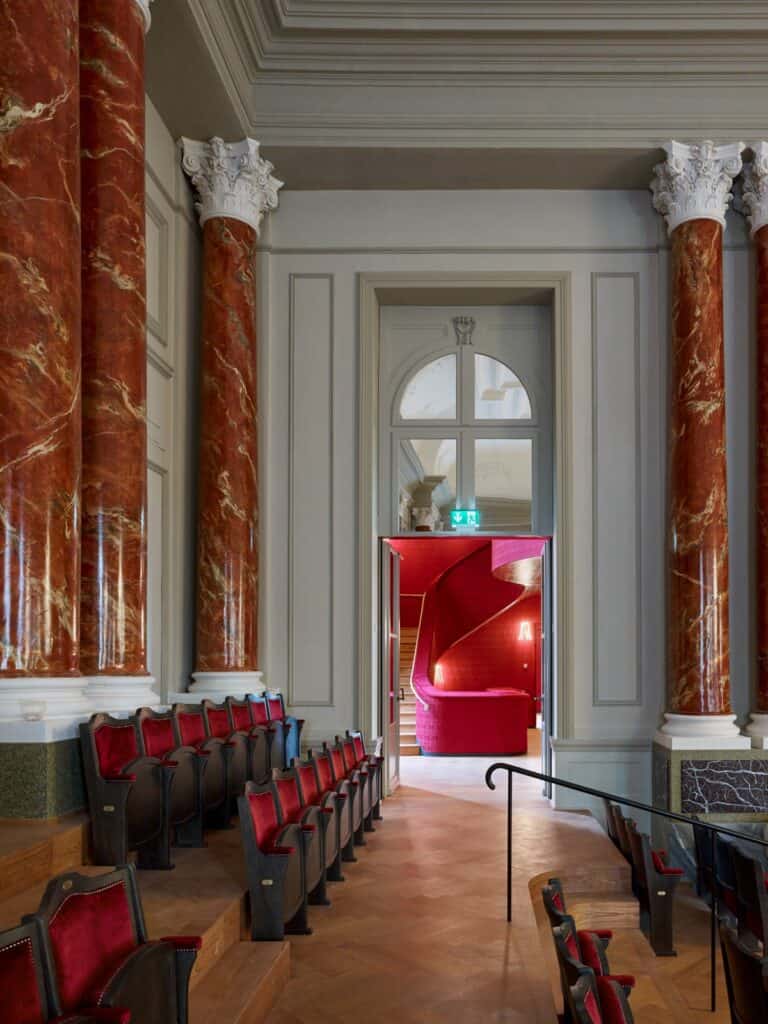 Aisle beside the stalls of the Musiksaal at the Stadtcasino Basel between marbled red columns with white Corinthian capitals, red velvet seats and herringbone parquet leading to a doorway onto a red staircase