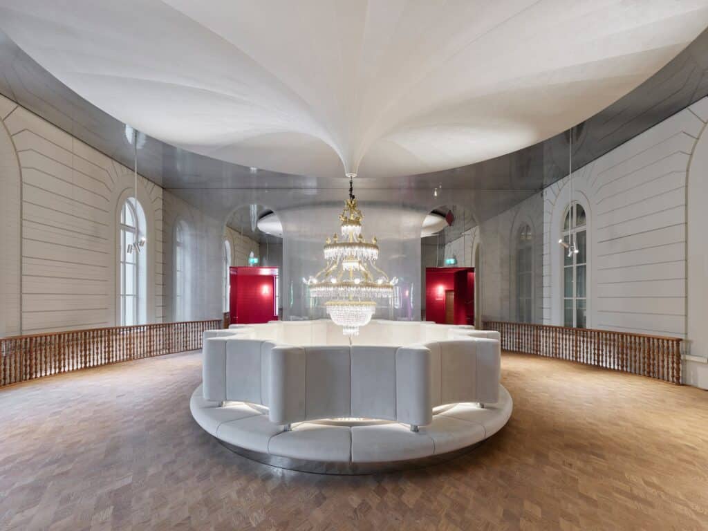 Upper foyer of the Stadtcasino Basel with a curving white upholstered balustrade ringing an opening in the floor beneath a draped white ceiling, a crystal chandelier, timber railings and arched windows in the old facade