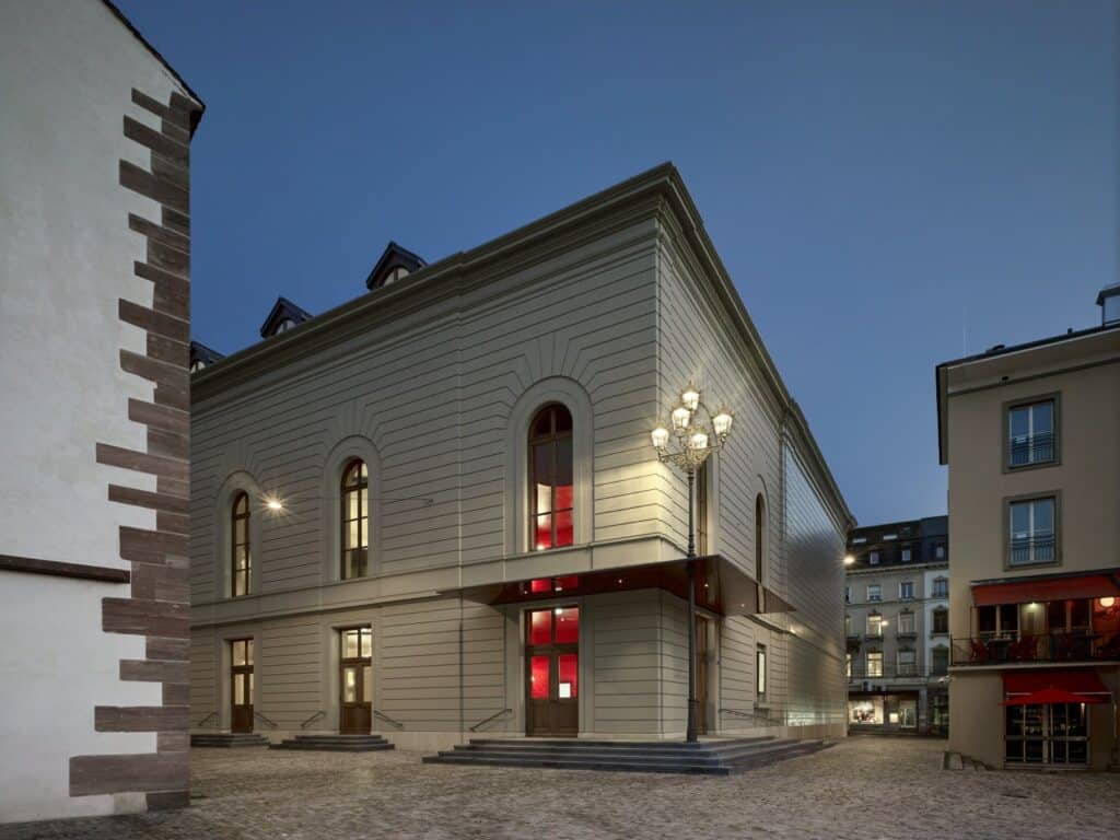 Corner of the Stadtcasino Basel extension at dusk, its pale stone-coloured facade with arched windows, a red-lit entrance under a canopy and a lantern-topped lamp post, the sandstone quoins of the Barfüsserkirche at the left edge