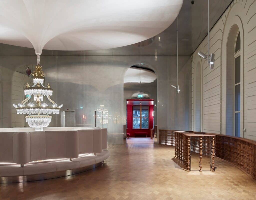 Crystal chandelier hanging from the draped white ceiling over a curving white balustrade in the upper foyer of the Stadtcasino Basel, a mirrored end wall, herringbone parquet and an old timber railing leading to a red doorway