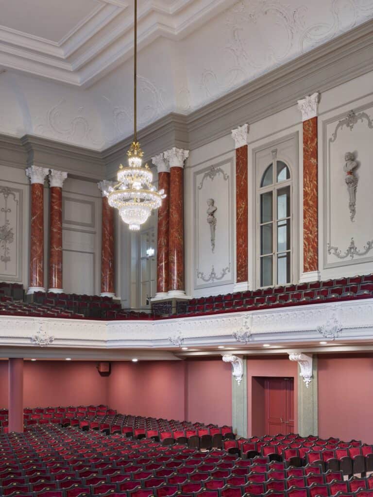 Tall view of the Musiksaal of the Stadtcasino Basel with a crystal chandelier before marbled red columns, stucco busts and an arched window above the balcony, rows of red seats in the stalls below