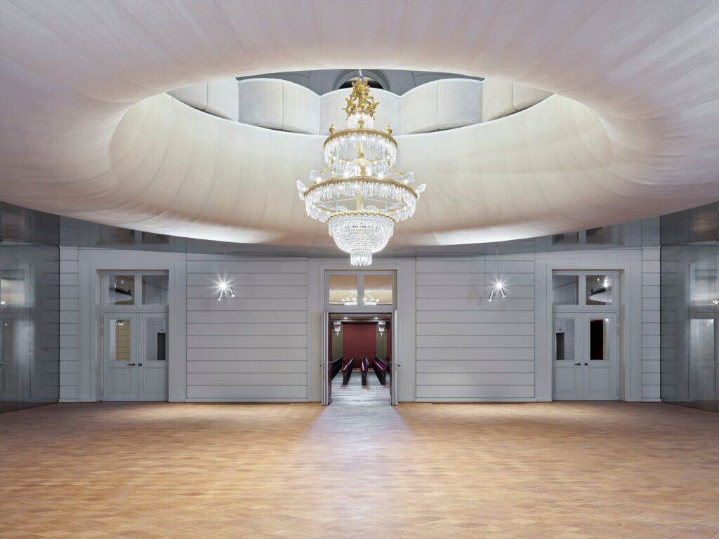 Ground floor foyer of the Stadtcasino Basel with oak parquet, grey panelled walls and doors open to the red-seated Musiksaal, a crystal chandelier hanging through the elliptical opening in the ceiling above
