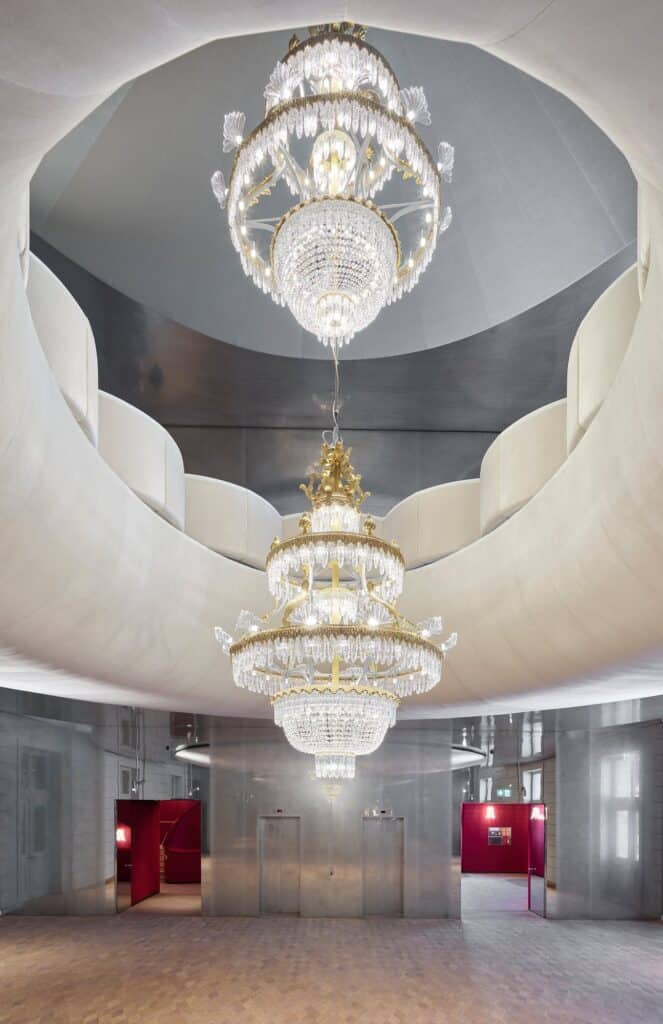Two crystal chandeliers hanging one above the other through the elliptical opening between the foyers of the Stadtcasino Basel, a curving white balustrade above and red doorways in the grey walls below