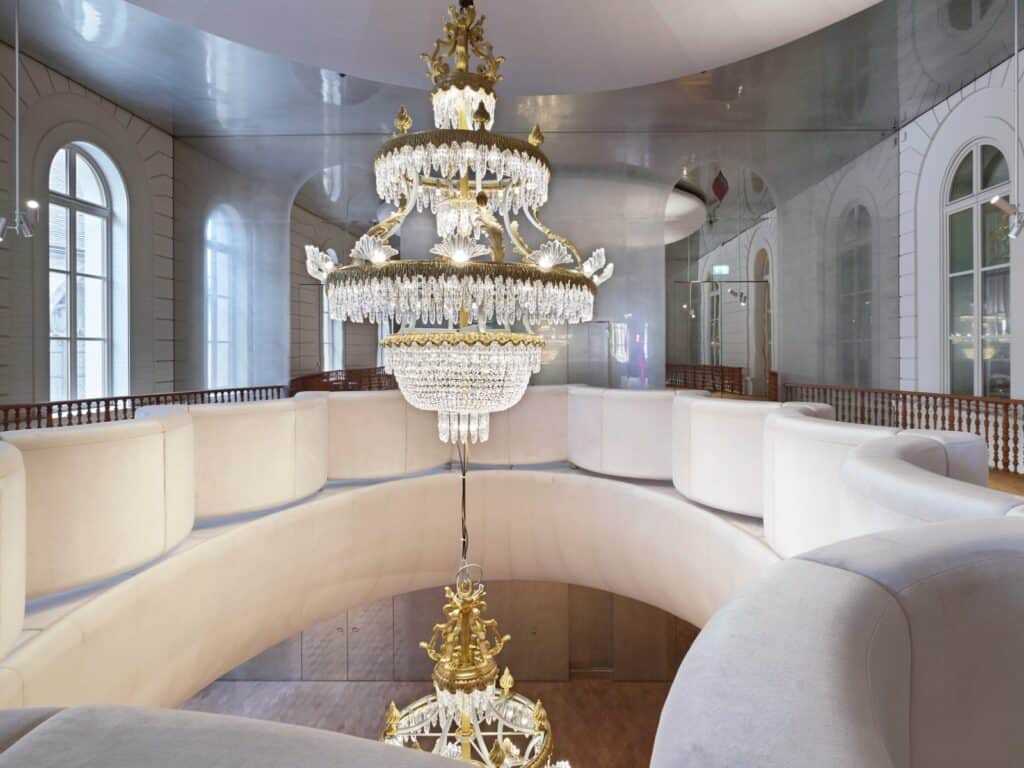 Gilded crystal chandelier hanging through the oval opening of the upper foyer at the Stadtcasino Basel, ringed by a curving white upholstered balustrade, a second chandelier below and arched windows reflected in mirrored walls