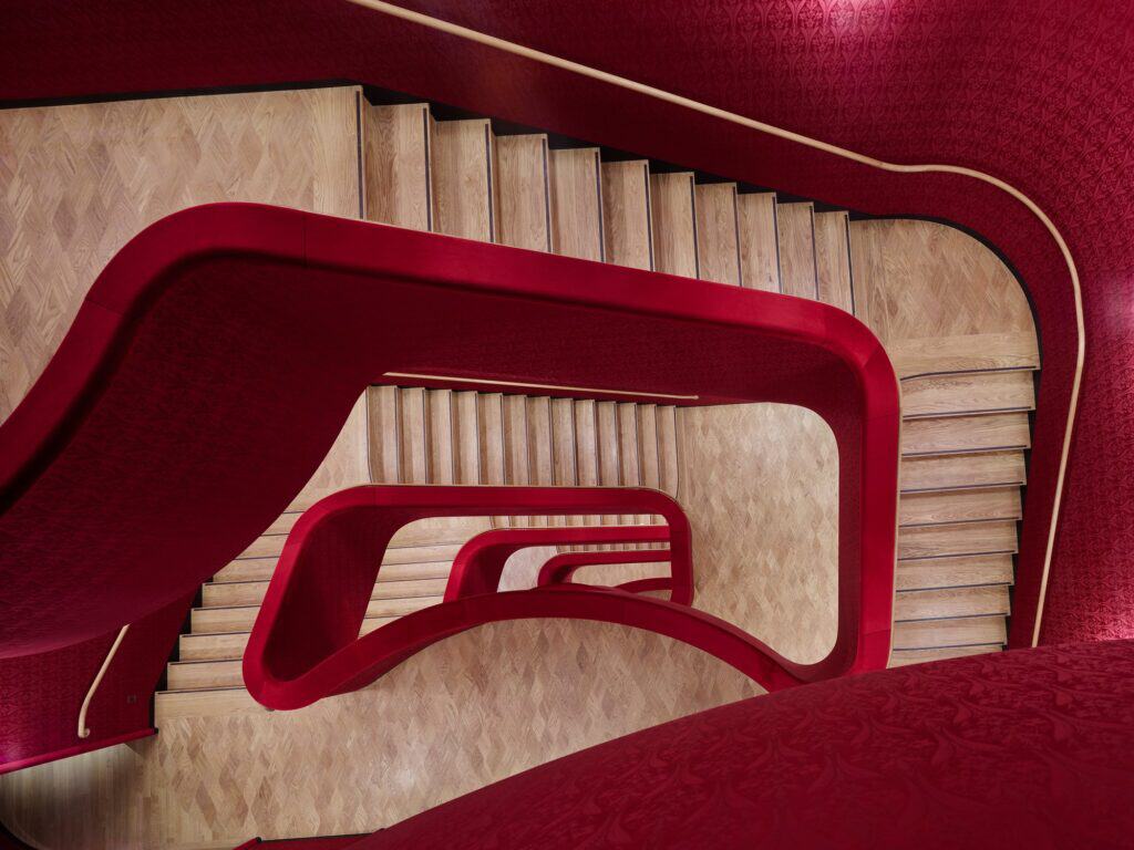 Looking down a staircase of the Stadtcasino Basel, curving red balustrades with brass handrails winding around flights of pale oak stairs and lens-patterned parquet
