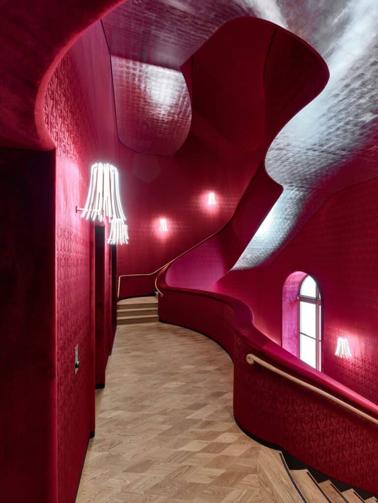 Curving staircase landing at the Stadtcasino Basel lined in red brocade, with fringed Parrucca wall lights, a small arched window and a silver-leafed ceiling above pale oak parquet