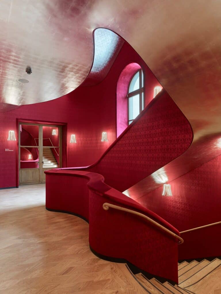 Red brocade staircase hall at the Stadtcasino Basel with a sweeping red balustrade and brass handrail, fringed Parrucca wall lights, an arched window and a shimmering silver metal-leaf ceiling over oak parquet