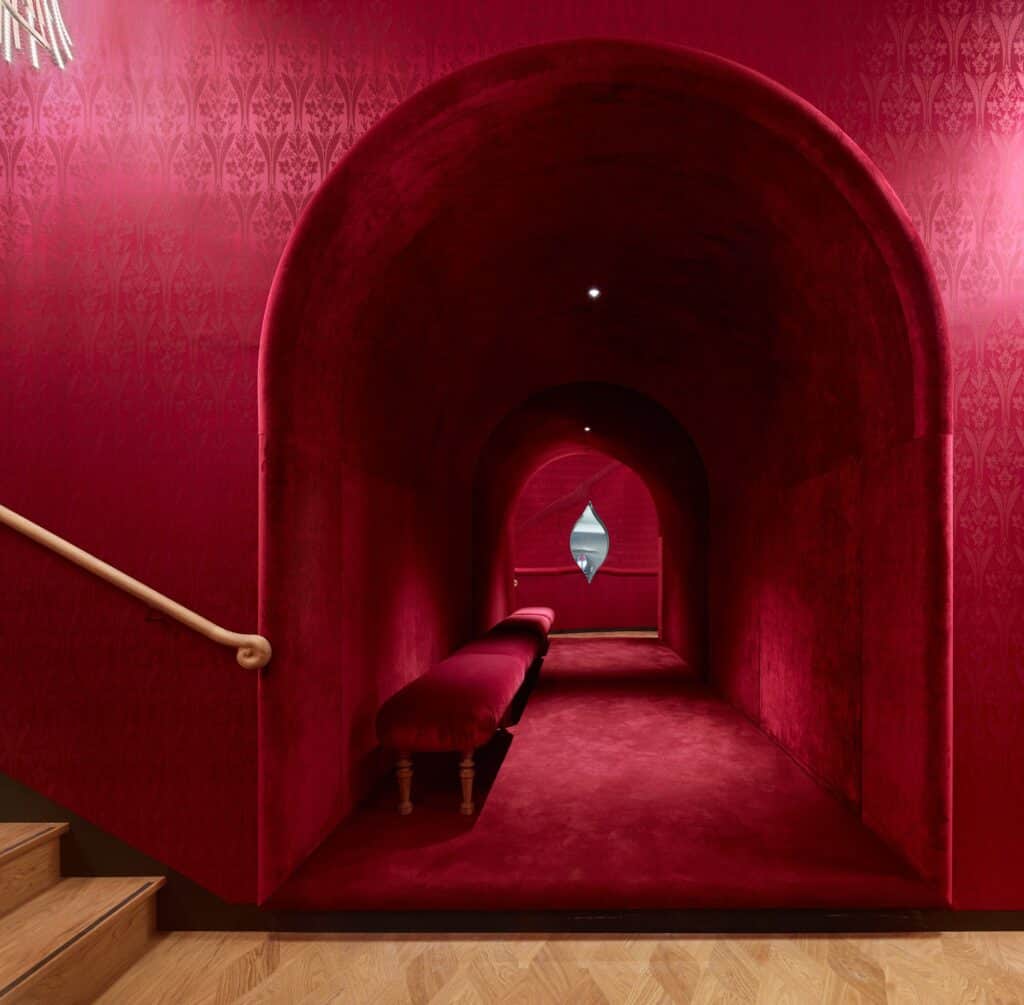 Arched loggia recess off a staircase at the Stadtcasino Basel upholstered in deep red velvet, with a red bench, receding arches and a small lens-shaped mirror at the end, a brass handrail on the red brocade wall