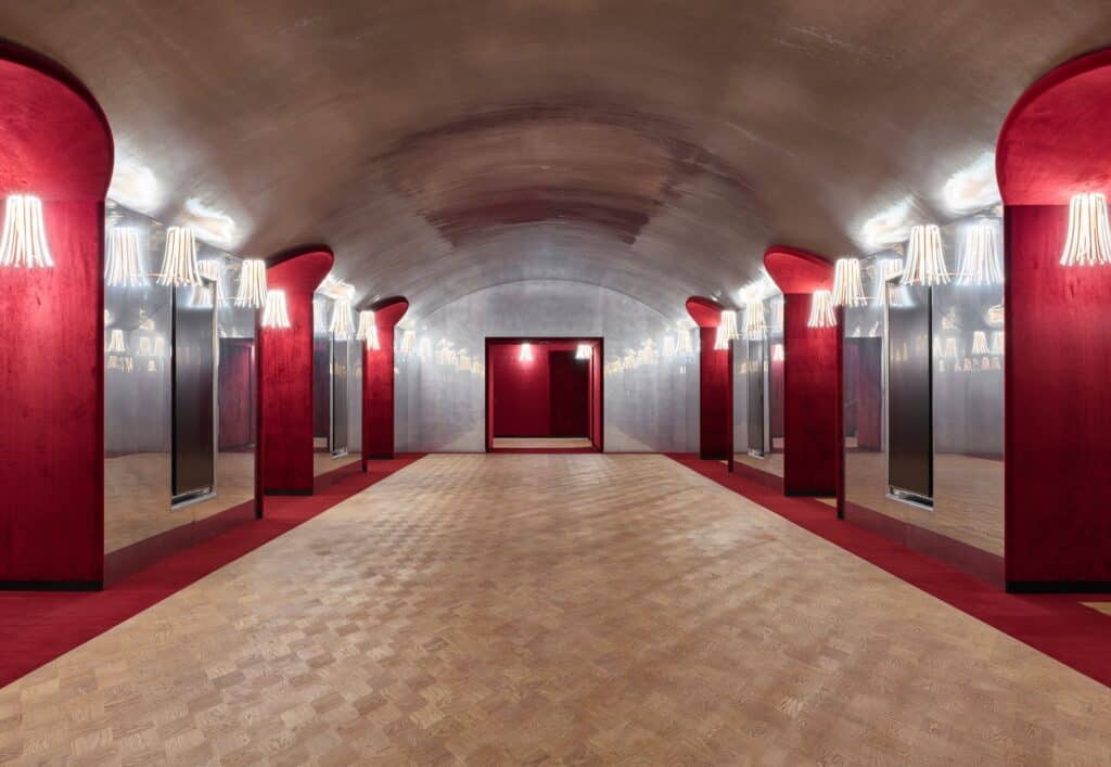 Vaulted lower foyer of the Stadtcasino Basel with a silvery barrel ceiling, red velvet-lined arched niches with fringed Parrucca lights and mirrors, and oak parquet leading to a red doorway