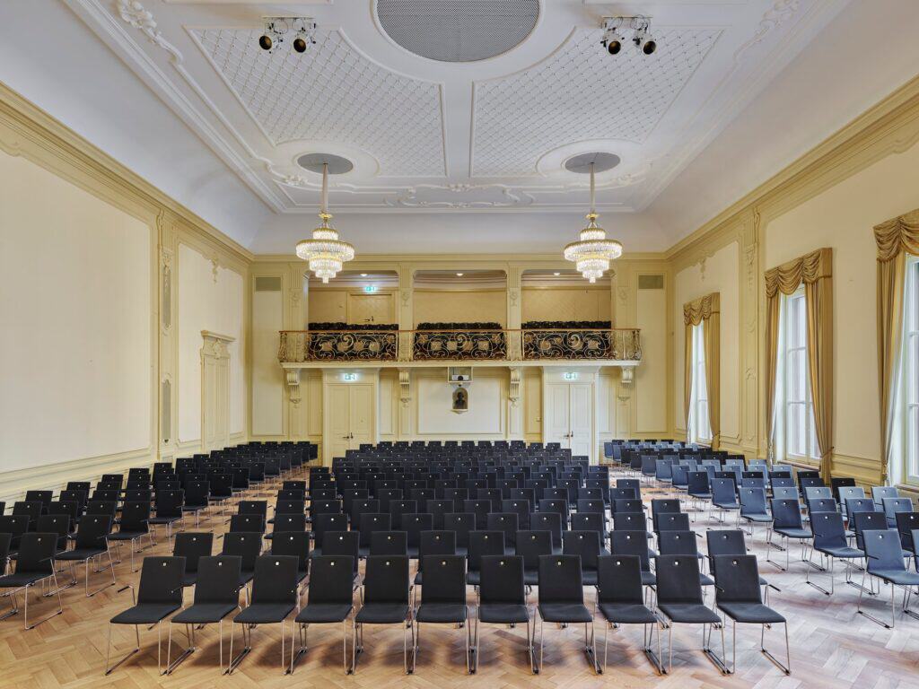 Hans Huber Saal at the Stadtcasino Basel with rows of dark chairs on herringbone parquet, pale yellow walls with gilt-trimmed pilasters, draped windows, a white stucco ceiling and a gallery with an ornate balustrade