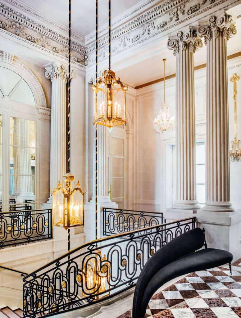 Listed 19th-century staircase at Hôtel de Crillon with a looping black wrought-iron balustrade, brass lanterns on long chains, white fluted columns and a checkered marble floor with a black sofa