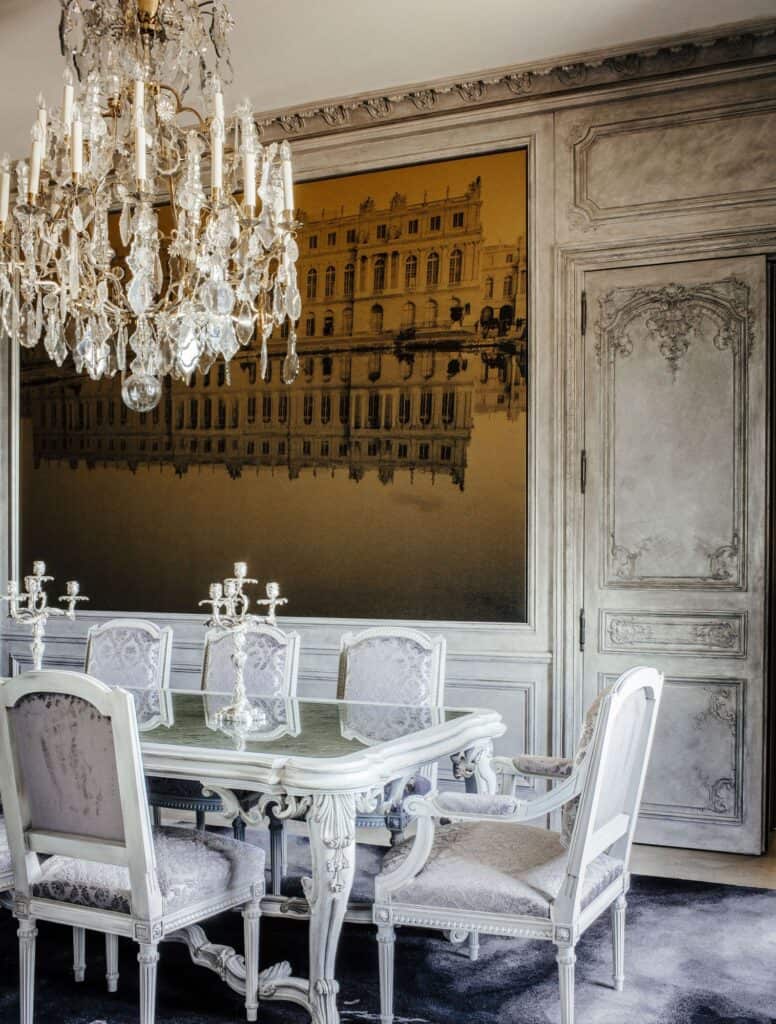 Dining room at Hôtel de Crillon with white carved Louis-style chairs around a marble-topped table, a crystal chandelier, grey painted boiserie and a large golden image of a palace on the wall