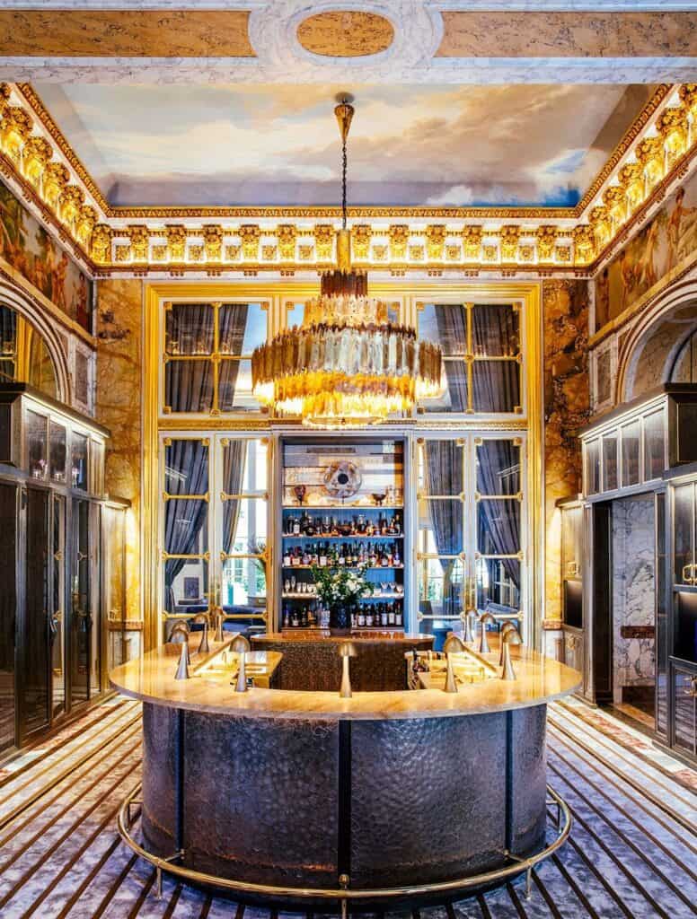 Horseshoe bar counter of Les Ambassadeurs at Hôtel de Crillon in dark hammered metal under a crystal chandelier, set within gilded walls of marble and mirrors on a striped carpet