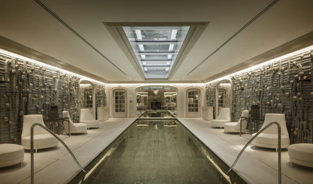 Underground pool at Hôtel de Crillon lit by a skylight, a long dark basin with steel handrails between white loungers and walls of textured gold and celadon ceramic