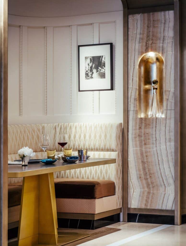 Dining banquette at Hôtel de Crillon with quilted cream and blush upholstery and a gold-based table, a backlit onyx panel with a gold wall light and a black and cream marble floor