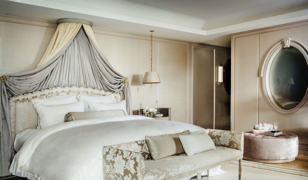 Guest room at Hôtel de Crillon with a white bed under a draped grey canopy, a carved bench with floral upholstery, an oval mirror and pale panelled walls
