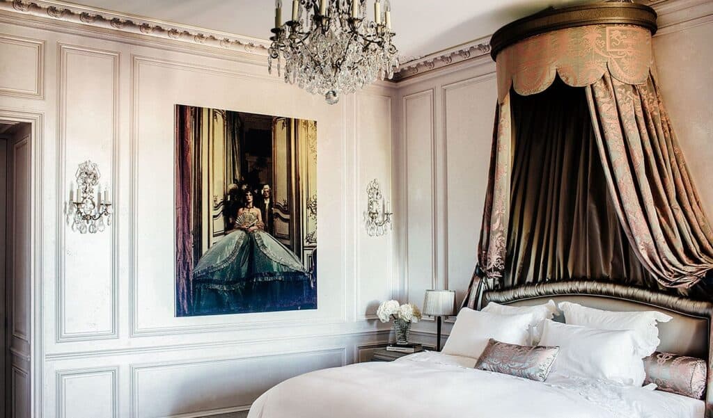 Bedroom at Hôtel de Crillon with a white bed beneath a gathered dark silk baldachin, white boiserie, a crystal chandelier and a large portrait of a woman in a ballgown