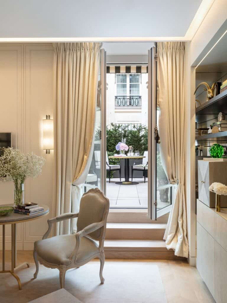 Sitting area of a Jardin suite at Hôtel de Crillon, a Louis-style armchair and side table with flowers beside cream curtains and French doors opening onto a private terrace