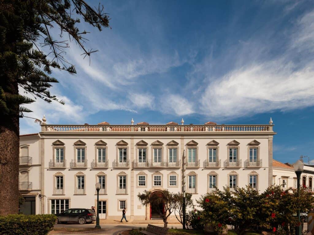 Hotel Palácio de Tavira, the white neoclassical facade of the former Palácio dos Tavares with stone-framed windows, wrought-iron balconies and a balustraded parapet with urns, beside a tall pine on the square