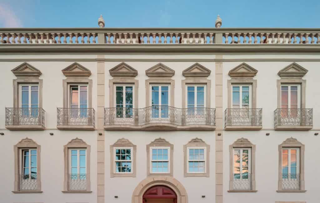 Restored main facade of Hotel Palácio de Tavira, pedimented stone window surrounds and wrought-iron balconies on a white wall divided by pilasters, above the arched red entrance door