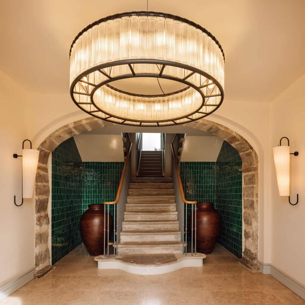 Entrance hall of Hotel Palácio de Tavira, a ring chandelier of glass rods before a stone arch framing the restored 18th-century stone staircase between deep green tiled walls and large clay jars