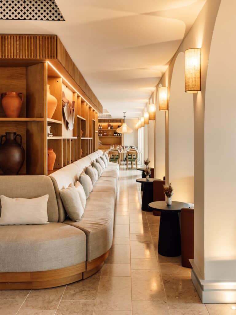 Lounge at Hotel Palácio de Tavira, a long curved banquette in pale linen beneath timber shelves of terracotta pots, with rattan wall lights along a white arcade and a stone floor