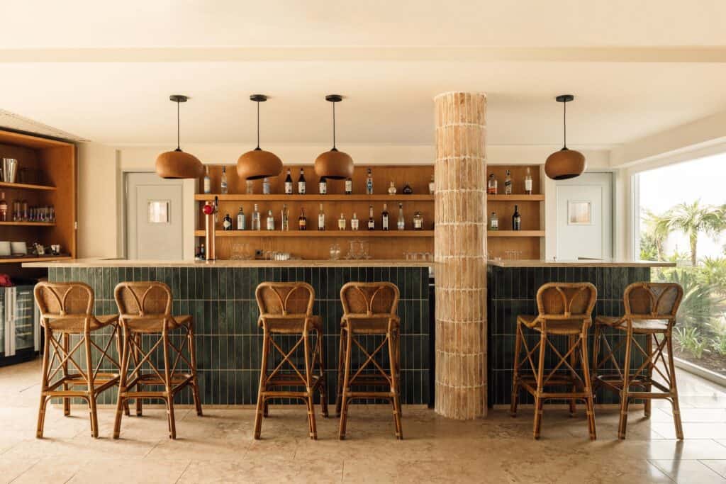 Bar at Hotel Palácio de Tavira, rattan bar stools before a counter clad in dark green tiles, copper pendant lamps, timber shelving of bottles and a rough stone column