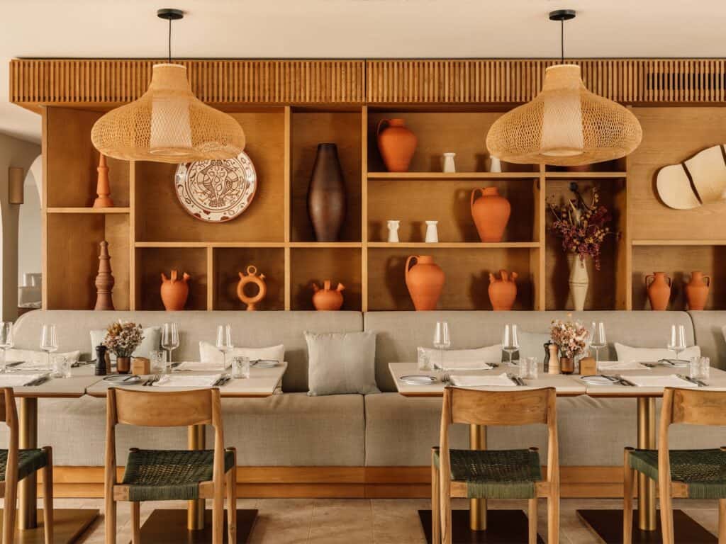Restaurant at Hotel Palácio de Tavira, set tables and timber chairs with green woven seats along a grey banquette, under woven rattan pendants and timber shelves of clay pots and plates