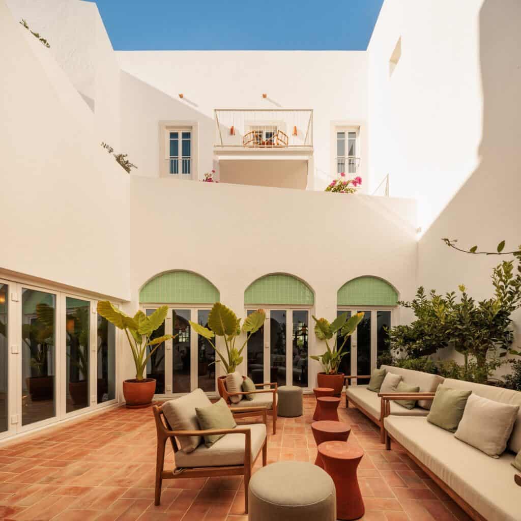 Courtyard terrace of Hotel Palácio de Tavira, timber sofas and armchairs with green cushions on terracotta tiles before glazed doors with green arched fanlights, enclosed by white walls