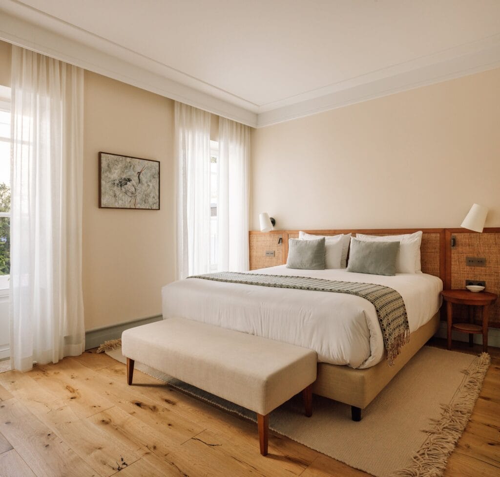 Guest room in the palace wing of Hotel Palácio de Tavira, a white bed with a striped throw and an upholstered bench on an oak floor, between tall windows with white linen curtains