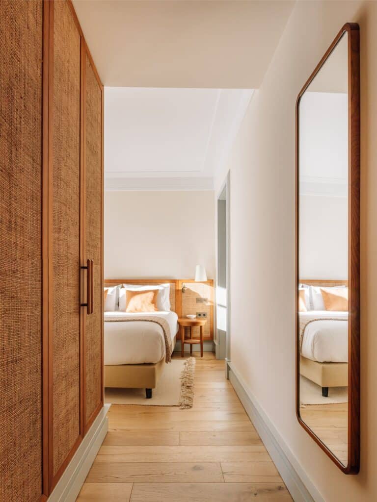 View through a guest room at Hotel Palácio de Tavira, past a rattan-fronted timber wardrobe and a tall timber-framed mirror along an oak-floored passage to the bed