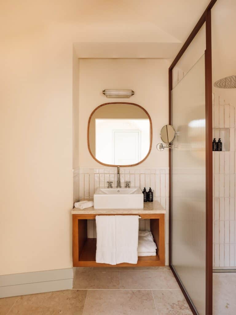 Bathroom at Hotel Palácio de Tavira, a white basin on a timber washstand beneath a rounded timber mirror, white tiles, a stone floor and a timber-framed glass screen
