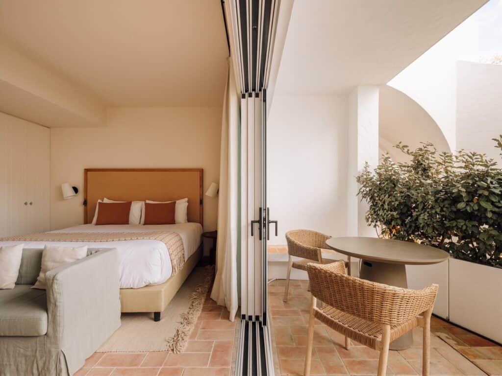 Medina guest room at Hotel Palácio de Tavira, a bed with terracotta cushions opening through folding glass doors onto a private terrace with rattan chairs, a round table and planters