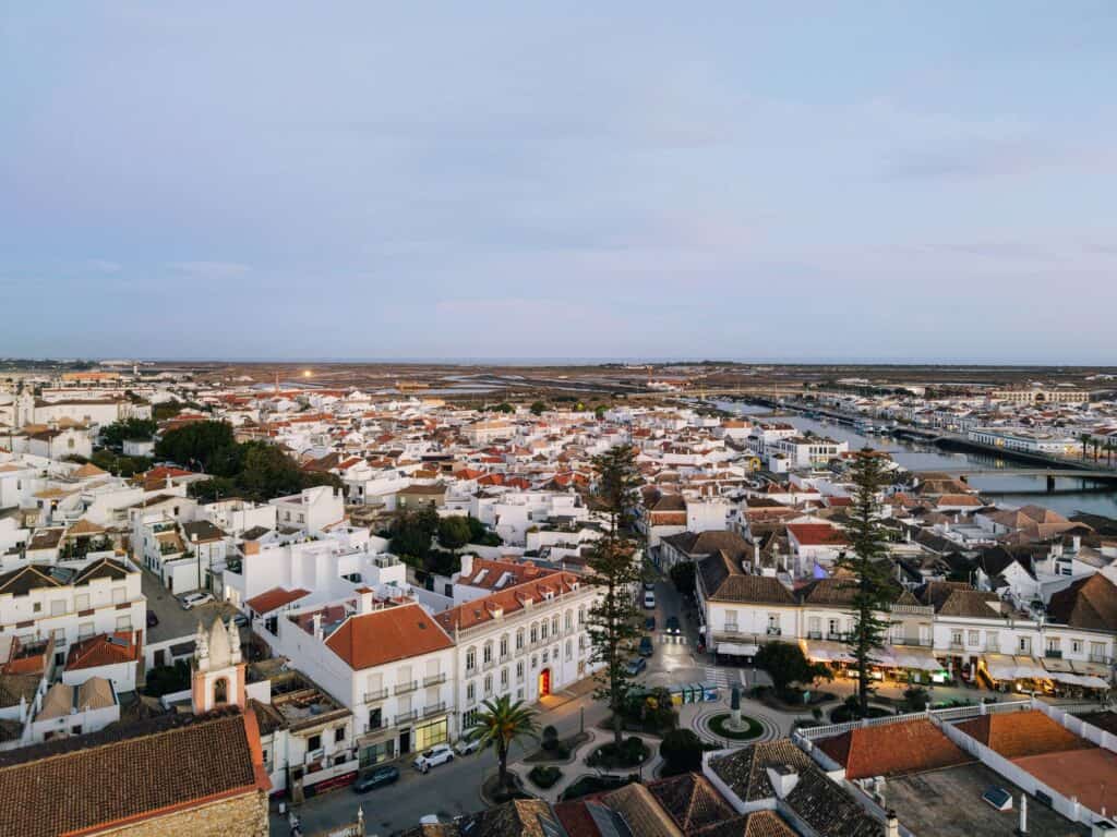 Aerial view of Hotel Palácio de Tavira in the historic centre of Tavira at dusk, the white palace on its square among red-tiled roofs, with the Gilão river and the Ria Formosa lagoon beyond