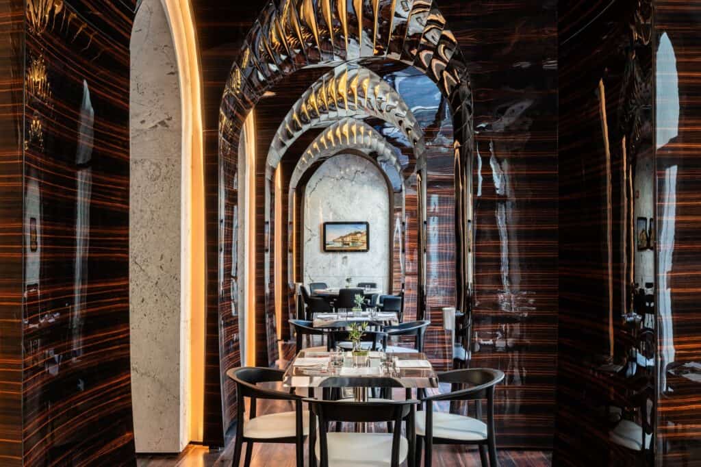 Bistro at Hotel Romeo Roma framed by glossy Makassar ebony walls, a sculpted vault of layered ribbed arches receding over dining tables with black chairs beside a pale stone arch