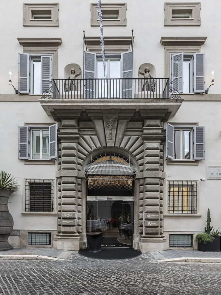 Facade of Palazzo Capponi on Via di Ripetta, home of Hotel Romeo Roma, with a rusticated stone portal and balcony, grey shutters and statues in niches above the cobbled street