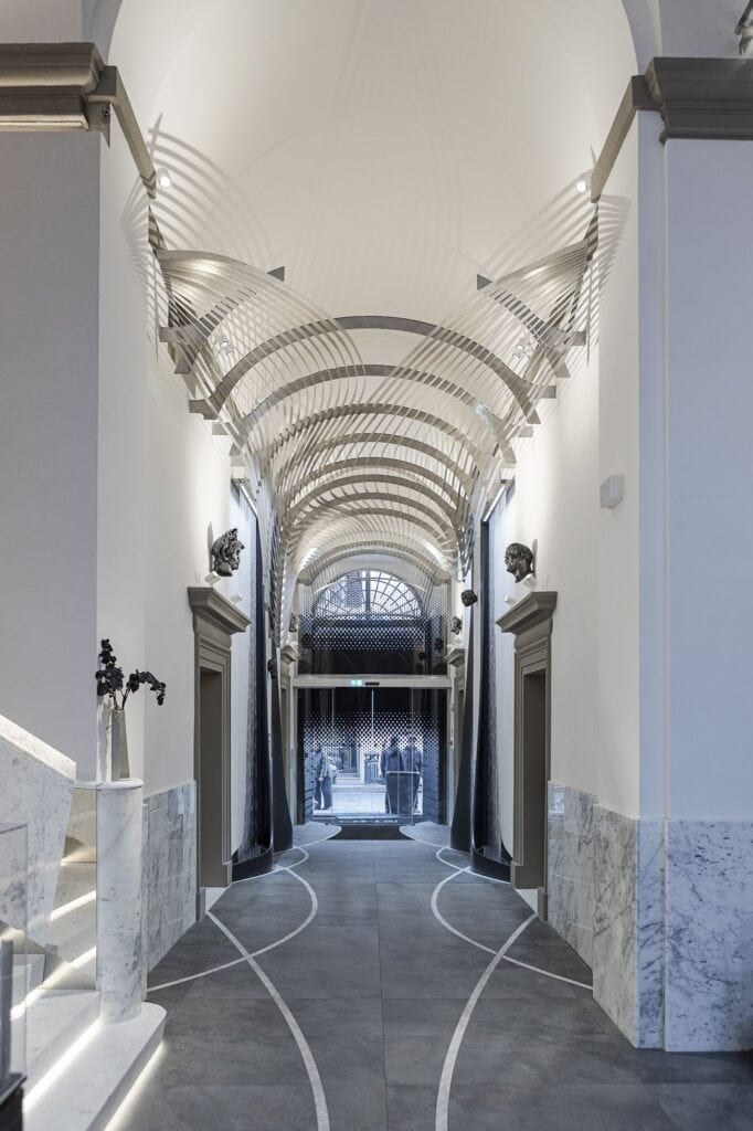 Vaulted white entrance passage of Hotel Romeo Roma lined with marble, a canopy of curved metal ribs overhead and inlaid lines sweeping across the dark stone floor towards the street gate