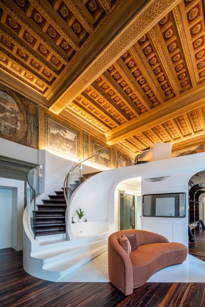 Reception at Hotel Romeo Roma beneath a gilded coffered Renaissance ceiling and frescoed walls, with a sculptural white staircase, a curved terracotta sofa and an ebony floor