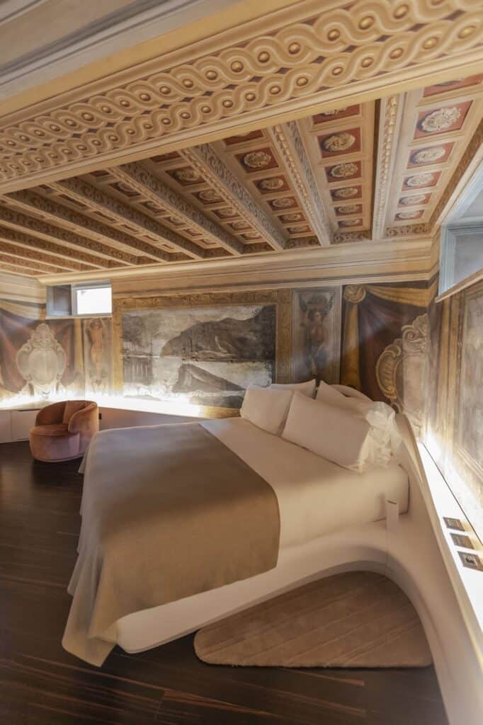 Bedroom at Hotel Romeo Roma under an ornate painted coffered ceiling, restored frescoes lit along the walls above a curved white bed platform on a dark ebony floor